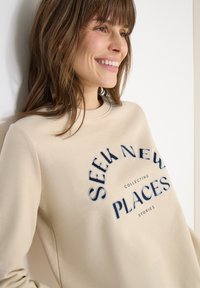 Femme souriante aux cheveux bruns portant un sweat-shirt beige avec le texte « Cherchez de nouveaux endroits, collectez des histoires » se tenant contre un mur blanc.