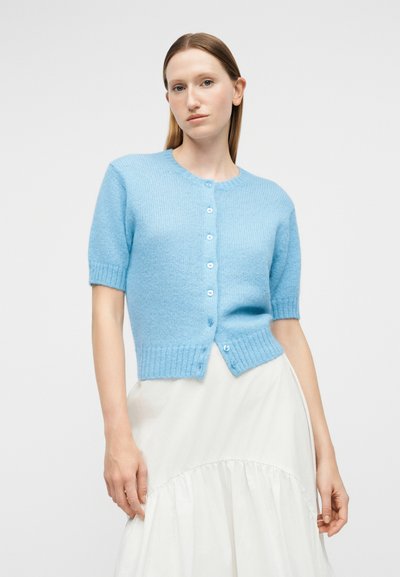 Femme portant un cardigan à manches courtes bleu clair boutonné et une jupe blanche taille haute sur un fond uni.