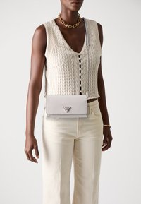 Sac bandoulière gris clair avec finition texturée, quincaillerie en argent et logo en triangle. Porté avec un haut en tricot crème et un pantalon beige clair.