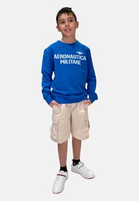 Felpa blu con stampa "AERONAUTICA MILITARE", pantaloni cargo beige e sneakers bianchi con dettagli rossi. Outfit casual sportivo.