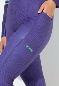 Fialové sportovní legíny s texturovaným zebrovým vzorem, vybavené malým zeleným logem "sports" a boční kapsou pro ukládání.