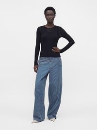 Femme debout, main sur la hanche, portant une chemise noire à manches longues, un pantalon large à rayures bleues, et des chaussures pointues de couleur claire sur fond blanc.