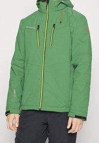 Giacca invernale verde con cappuccio e zip gialla, due tasche a soffietto con zip sul petto, polsini regolabili e pantaloni neri su una persona in piedi.