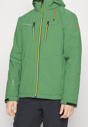 Giacca invernale verde con cappuccio e zip gialla, due tasche a soffietto con zip sul petto, polsini regolabili e pantaloni neri su una persona in piedi.