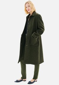 Manteau vert à motif pied-de-poule avec col, manches longues, boutons à l'avant et poches. Porté par-dessus un haut noir et un pantalon vert avec des ballerines noires.