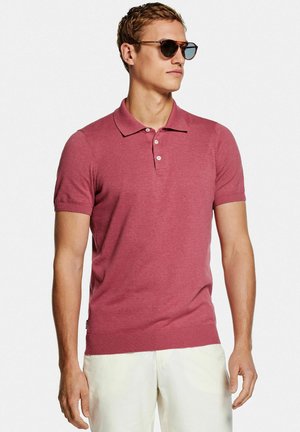 Uomo con occhiali da sole marroni, una polo rosa rossa a maniche corte e pantaloni bianchi in piedi davanti a uno sfondo uniforme.