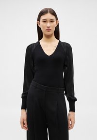 Sieviete ar taisniem tumšiem matiem, tērpusies melnā V apkakles topā ar zīdainām piedurknēm un melnās biksēs ar augstu jostasvietu, stāvot pret vienkāršu fona sienu.