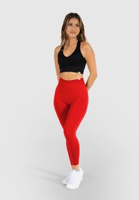 Rote hoch taillierte Leggings mit glatter Textur, kombiniert mit einem schwarzen, gerippten, kurz geschnittenen Tanktop. Modell trägt weiße Sportschuhe.