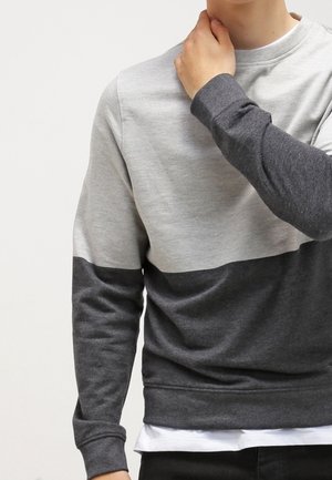 Persona che indossa una felpa grigia bicolore, mano poggiata sul collo, in piedi contro uno sfondo chiaro e uniforme.