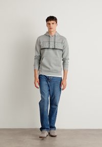 Gray hoodie with embossed logo, front kangaroo pocket, paired with blue denim jeans and gray sneakers, standing on a neutral floor.