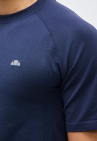 Κοντινό πλάνο ενός ατόμου που φοράει αθλητική μπλούζα με κοντά μανίκια σε ναυτικό μπλε χρώμα, με δίχτυ στα πλαϊνά και μικρό λευκό λογότυπο στο στήθος.