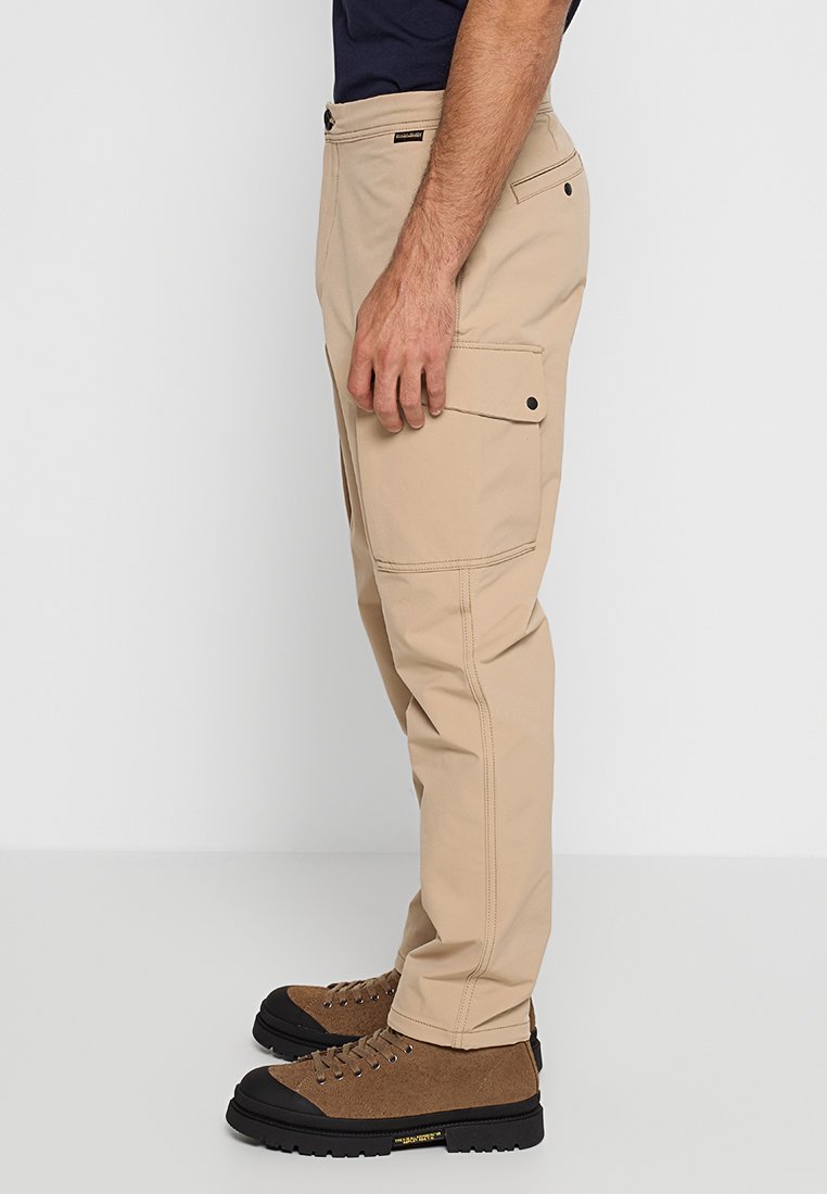 Pantaloni cargo beige con una vestibilità dritta, realizzati in tessuto resistente, dotati di tasche laterali e cuciture a contrasto. Indossati con scarponi da trekking marroni.
