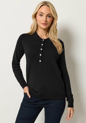 Blondynka ubrana w czarną koszulkę polo z długim rękawem z pięcioma białymi guzikami i ciemnoniebieskie dżinsy, stoi z jedną ręką w kieszeni.