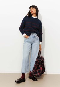 Pull marin avec des rayures bordeaux, jean large bleu clair, chaussures noires et sac à carreaux. Le col de la chemise blanche est visible sous le pull.