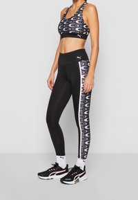 Kobieta w czarnym i wzorzastym stroju sportowym Puma, w legginsach i białych skarpetkach Puma, w czarnych butach sportowych, stojąca w pobliżu szarej ściany.