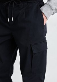 Black cargo pants with an elastic waistband and drawstring. Features side pockets and a flap pocket. Material appears smooth and durable.