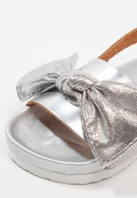 Srebrny sandał z dużą kokardą, metaliczne wykończenie, otwarty dziób, fakturowana cholewka i wyściełana podeszwa. Regulowany pasek na kostkę niewidoczny.