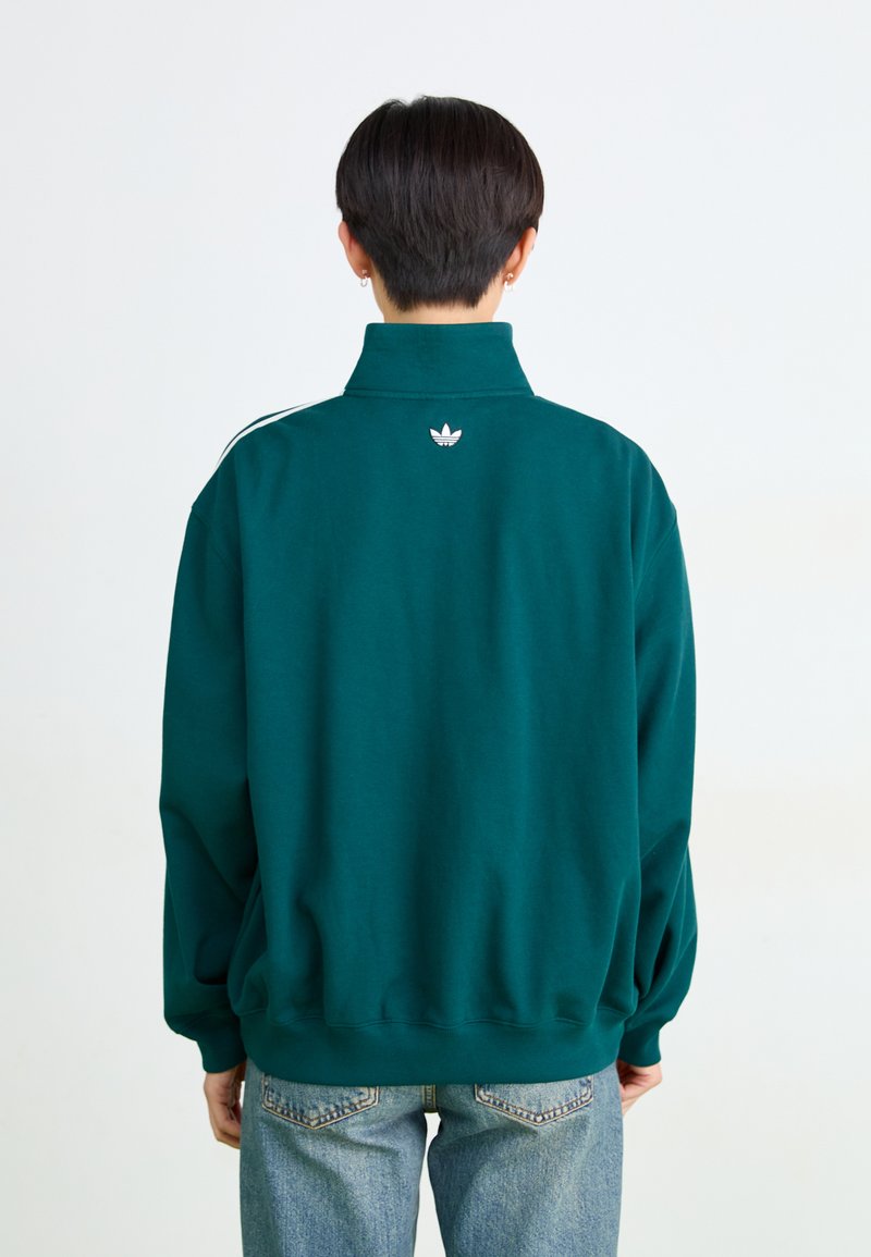Sweatshirt oversize vert foncé avec un col haut, rayures blanches sur les épaules et un petit logo blanc à l'arrière. Tissu doux et coupe décontractée.