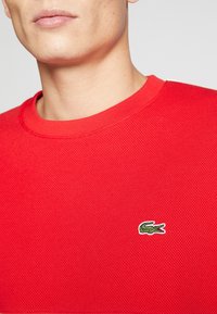 Czerwona strukturalna sweter z okrągłym dekoltem i krótkimi rękawami, z haftowanym zielonym logo krokodyla na piersi. Materiał z domieszką bawełny.