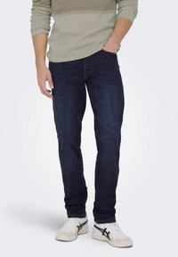 Mörkblå denimjeans med rak passform, kombinerat med en ljusgrå texturerad långärmad skjorta och vita sneakers med svarta detaljer.