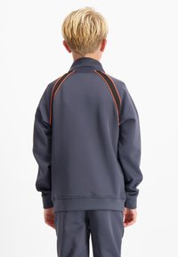 Blond persoon die een donkergrijze trainingspak met oranje bies draagt, staande met de rug naar de camera tegen een effen witte achtergrond.