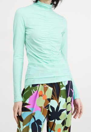 Henkilö, joka pukeutunut vaaleanturkoosiin röyhelöityyn pitkähihnaiseen yläosaan ja monivärisiin trooppisiin kuositettuihin housuihin, joissa on vihreitä, purppuran ja oranssin sävyjä.