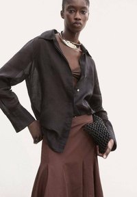 Schwarzes transparentes Langarmshirt über einem braunen Oberteil und einem taillierten braunen Faltenrock. Hält eine kleine schwarze geflochtene Clutch. Silberne Halskette als Akzent.