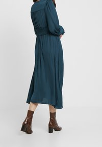 Frau in einem türkisfarbenen Midi-Kleid mit langen Ärmeln und braunen strukturierten Stiefeletten mit Absatz, steht vor einem schlichten hellen Hintergrund.