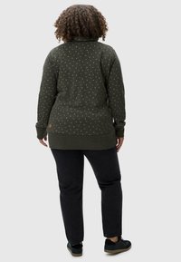 Suéter verde oscuro con pequeños patrones blancos, mangas largas y puños acanalados. Combinado con jeans oscuros y zapatillas negras.