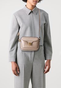 Béžová kožená crossbody taška se zlatou sponou, obdélníkového tvaru, hladkého povrchu a ramenním popruhem, nošena přes oblečení s kostkovaným vzorem.