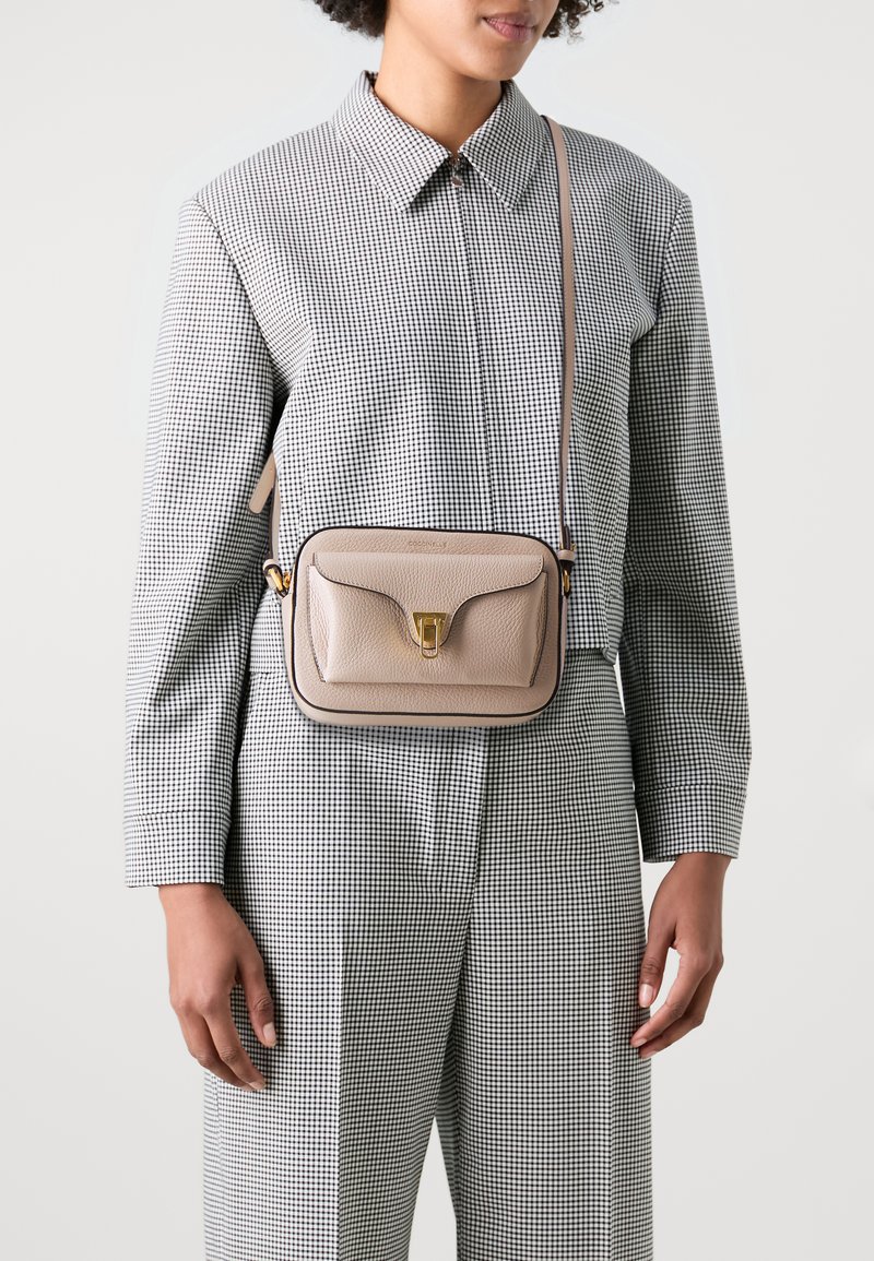 Béžová kožená crossbody taška se zlatou sponou, obdélníkového tvaru, hladkého povrchu a ramenním popruhem, nošena přes oblečení s kostkovaným vzorem.