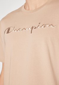 Hellbeige Baumwoll-T-Shirt mit einem glänzenden, geprägten "Champion"-Logo in einer kursiven Schriftart quer über die Brust. Kurze Ärmel und Rundhalsausschnitt.