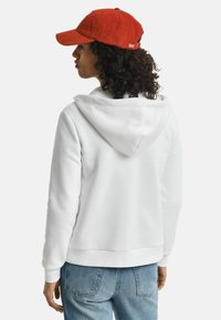 Witte hoodie met een capuchon, ribbel manchetten en zoom. Gepaard met een rode fluwelen pet. Blauwe spijkerbroek zichtbaar. Achteraanzicht.