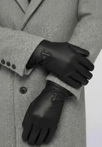 Gants en cuir noir avec quatre coutures et un logo ton sur ton, portés sur un manteau gris en chevrons avec trois boutons et un col cranté.