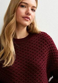 Jonge vrouw met lang blond haar, gekleed in een gestructureerde donkerrode gebreide trui, recht naar voren kijkend met een neutrale uitdrukking tegen een lichte achtergrond.