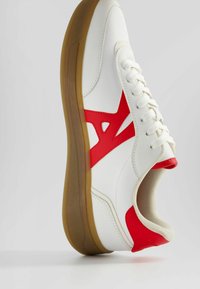 Vit sneaker med röda accenter, slät syntetisk ovandel, rund tå och texturerad beige gummisula. Har snörning och logotypdetalj på sidan.
