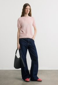 Γυναίκα που φοράει ροζ πουλόβερ με κοντά μανίκια, σκούρο τζιν φαρδιάς γραμμής, ροζ παπούτσια, κρατώντας με το ένα χέρι μαύρη τσάντα με πιέτες, στέκεται σε γκρι πάτωμα.