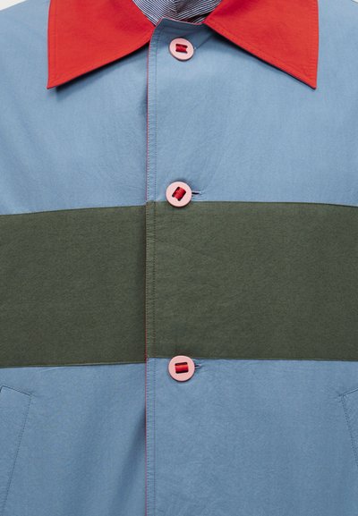 Πουκάμισο με κόκκινο γιακά, ανοιχτό μπλε άνω και κάτω τμήματα, φαρδιά οριζόντια χακί ρίγα και ροζ κουμπιά με κόκκινη κλωστή.