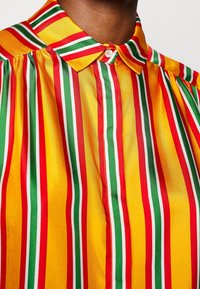 Gros plan sur un col de chemise à rayures verticales rouges, jaunes, vertes et blanches sur un tissu brillant, porté par une personne à la peau foncée.