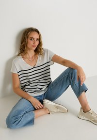 Camiseta gris de cuello en V con rayas horizontales en azul marino, combinada con jeans de mezclilla azul claro. El modelo está sentado de manera informal sobre una superficie blanca.