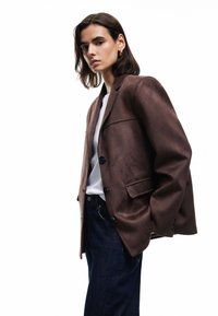 Brauner Wildlederblazer mit tailliertem Schnitt, verfügt über zwei Knöpfe, eine einzelne Brusttasche und eine leicht strukturierte Oberfläche. Getragen über einem weißen T-Shirt.