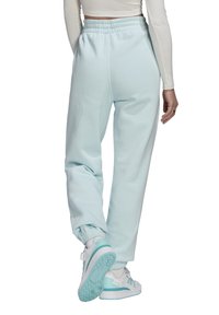 Kobieta ubrana w jasnoniebieskie dresy i białe sneakersy, stojąca z jedną nogą lekko uniesioną, pokazująca widok z tyłu.