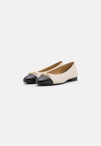 Ballet flats vaaleassa nahassa, jossa on kiiltävä musta lakatun kärjen. Edessä on kultaketjusta tehty yksityiskohta. Litteä pohja, minimalistikko muotoilu.