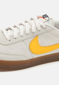 Šedé semišové tenisky s žlutým logem Nike, bílými tkaničkami a hnědou gumovou podrážkou, s texturovaným povrchem a prošitými detaily.