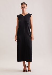 Robe longue noire sans manches avec un décolleté en V, texture lisse, coupe droite et une couture centrale. Associée à des talons noirs à lanières.