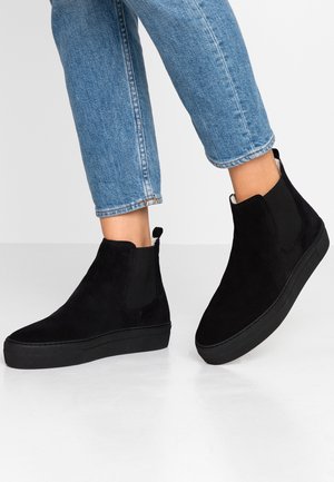 Bottines en daim noir avec languettes, portées avec un jean bleu retroussé sur un fond clair uni.