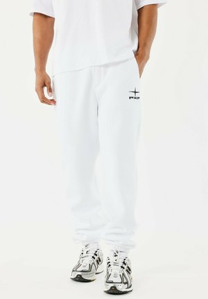 Uomo che indossa pantaloni sportivi bianchi con il logo PXP sulla coscia, abbinati a scarpe da corsa bianche e nere, in piedi su uno sfondo semplice.