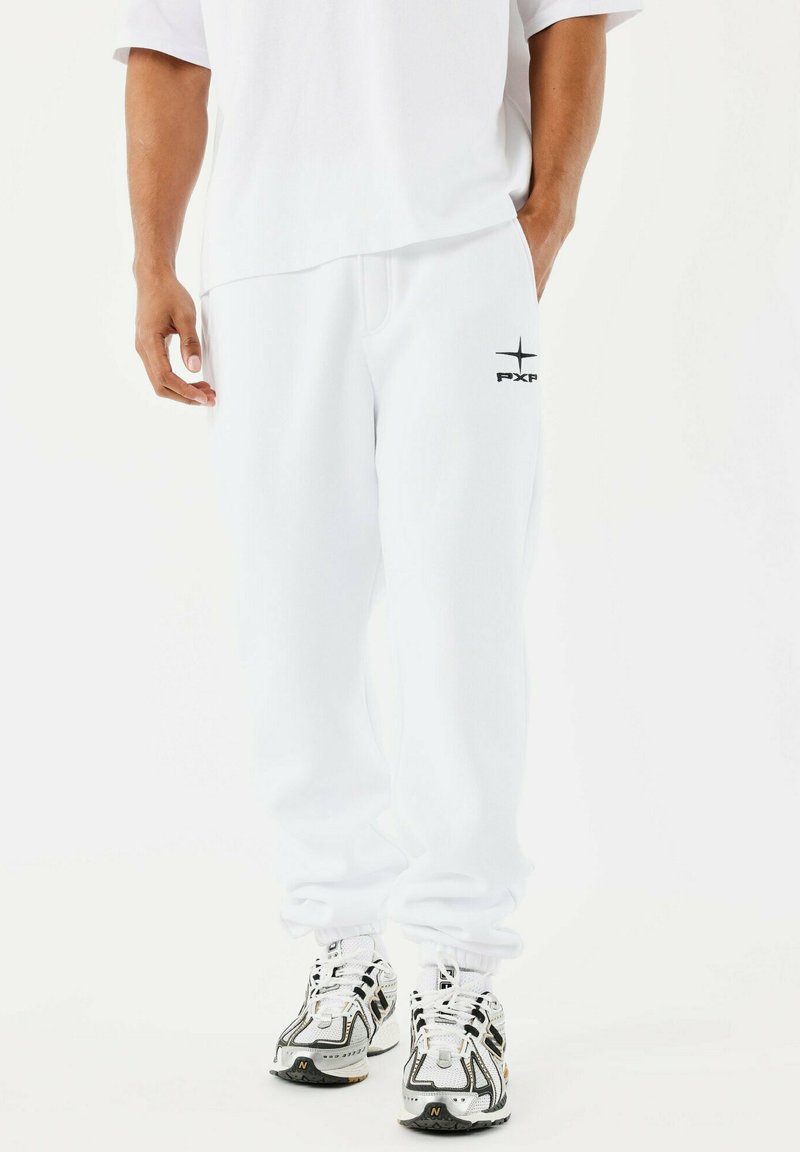 Man in witte sweatpants met PXP-logo op de dij, gecombineerd met wit en zwarte hardloopschoenen, staande tegen een effen achtergrond.