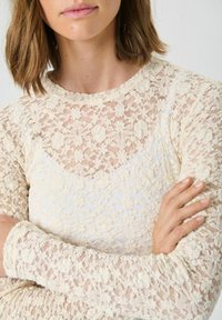 Top din dantelă în crem, cu mâneci lungi și un decolteu înalt. Țesătura prezintă un model floral cu un aspect texturat și transparent.