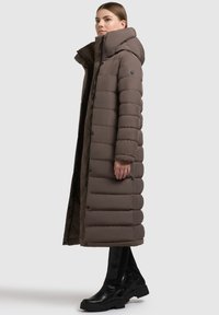 Lang brun pufferjakke med hætte, der har et quiltet design, trykknapper og en slank silhuet. Bæres med sorte læder ankelstøvler.